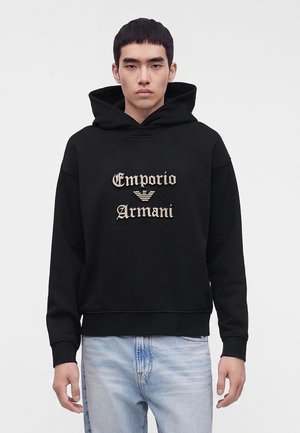 Νέος άνδρας που φοράει μαύρο φούτερ Emporio Armani με μπεζ λογότυπο και ανοιχτό μπλε τζιν, στέκεται μπροστά σε ένα απλό λευκό φόντο.