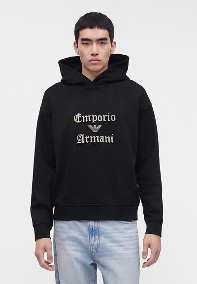 Млад мъж, носещ черен суичър Emporio Armani с бежов логотип и светлосини дънки, изправен пред бял фон.