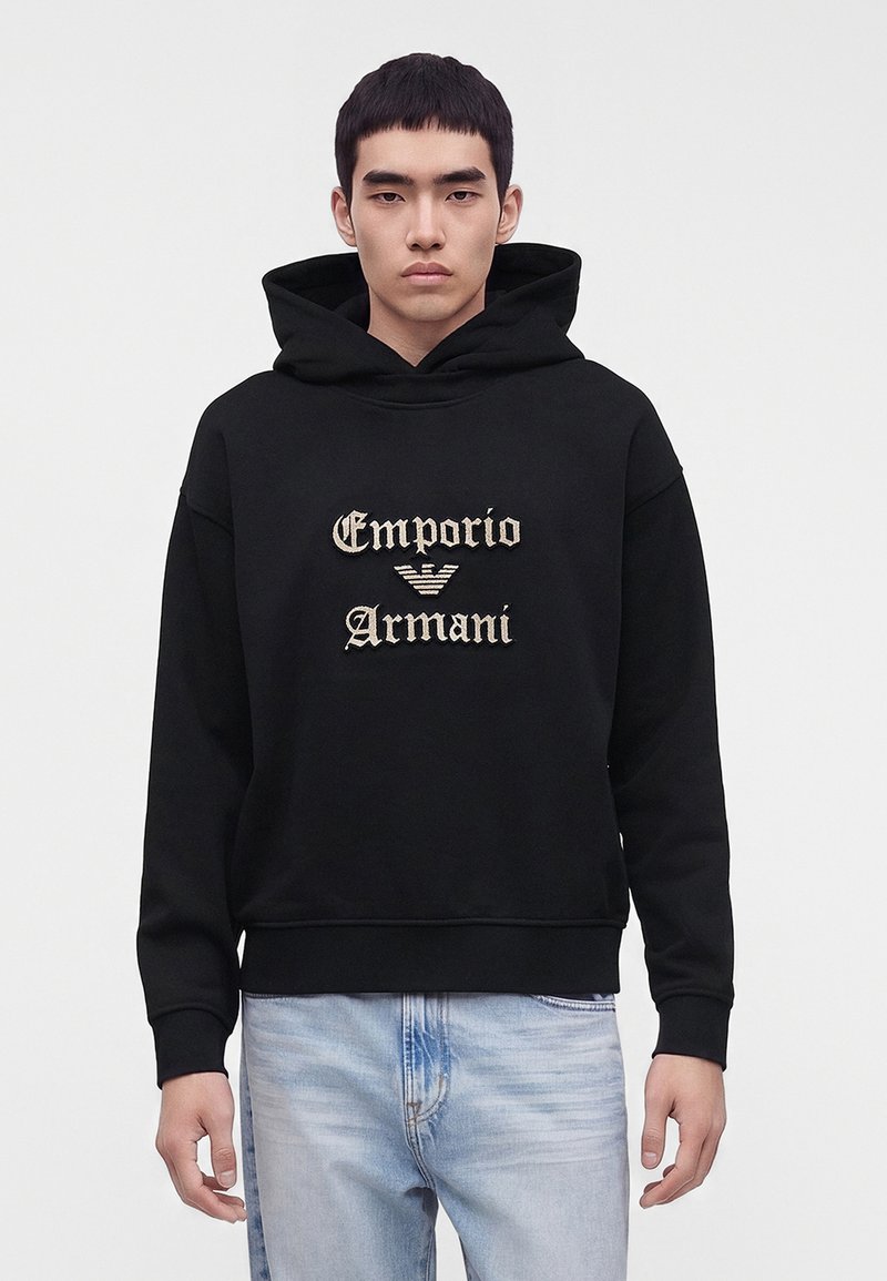 Νέος άνδρας που φοράει μαύρο φούτερ Emporio Armani με μπεζ λογότυπο και ανοιχτό μπλε τζιν, στέκεται μπροστά σε ένα απλό λευκό φόντο.