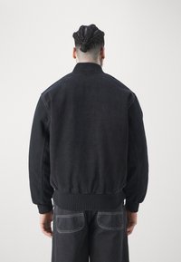 Czarna kurtka bomberka z denimu z prążkowanym kołnierzem i mankietami na nadgarstkach. Charakteryzuje się gładką fakturą oraz elastycznym pasem w talii. Widok z tyłu ukazuje prosty design.