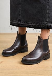Bottes chevilles en cuir marron avec des panneaux élastiques sur les côtés, bout rond et semelle texturée, associées à une jupe en denim noir effilochée.