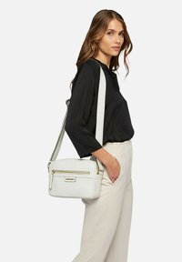 Borsa a tracolla bianca realizzata in materiale testurizzato, con una zip frontale e dettagli in hardware dorato. Le cinghie sono regolabili.