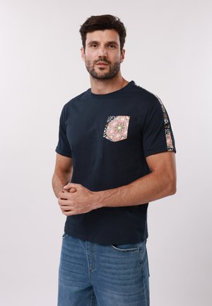 Άνδρας με μπλε ναυτικό μπλουζάκι με τσέπη και ρίγα στο μανίκι με σχέδιο, στέκεται με τα χέρια σταυρωμένα και κοιτάζει την κάμερα.