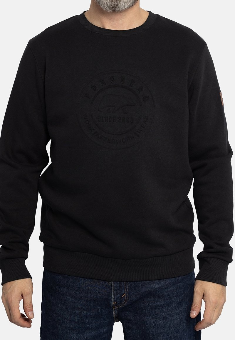 Homme portant un sweat-shirt noir à manches longues avec un logo circulaire subtilement embossé et un jean bleu foncé sur fond blanc.