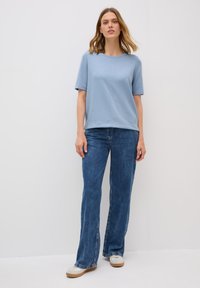 Vrouw staat zelfverzekerd, gekleed in een lichtblauw shirt en een wijde denim jeans, tegen een effen witte achtergrond.