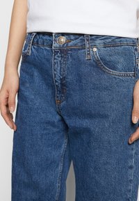 Personne portant un jean taille haute bleu en denim avec un bouton en métal et des coutures orange, ainsi qu'une chemise blanche rentrée dedans.