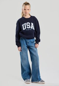 Navy sweatshirt with "USA New York City" print, oversized design. Wide-leg light blue jeans, faded denim texture, casual sneakers.
