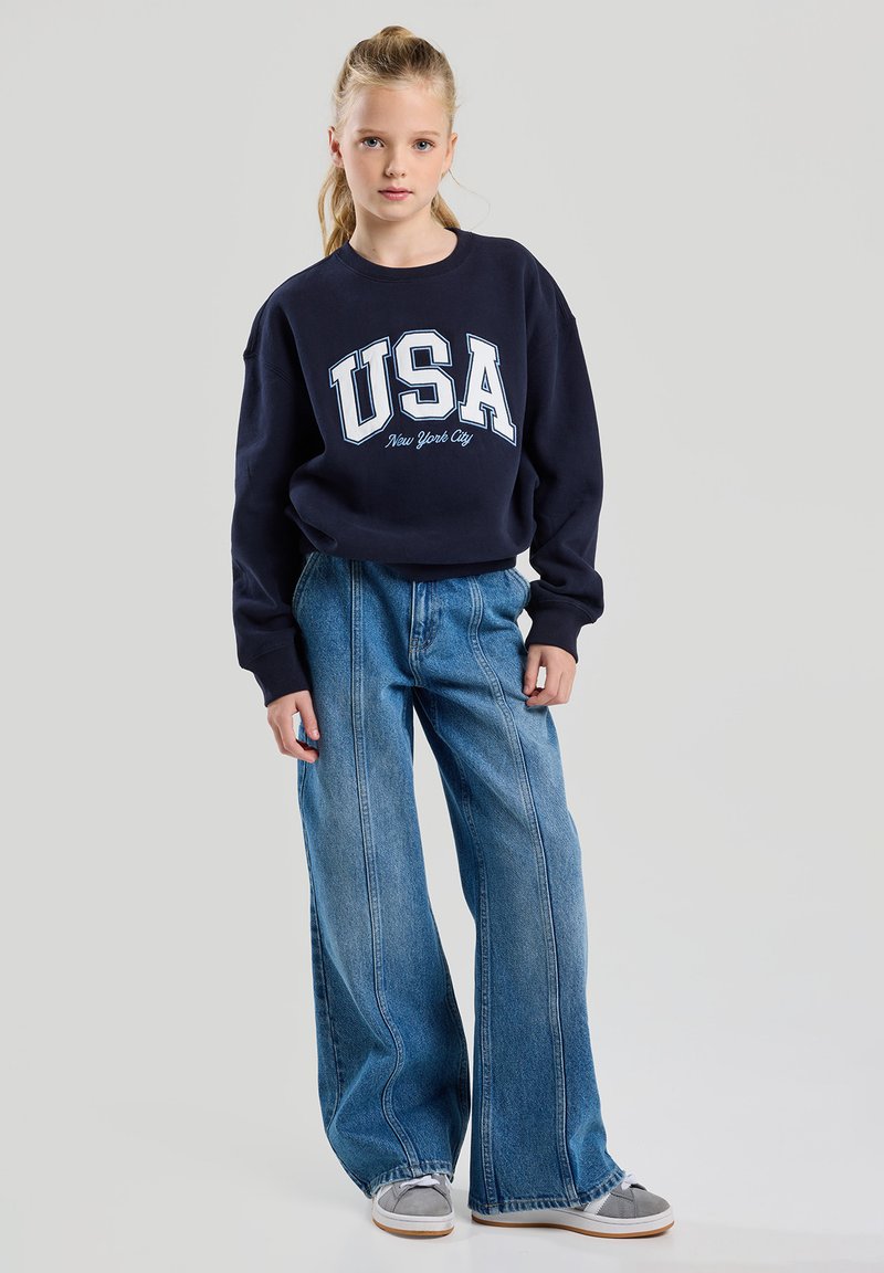 Navy sweatshirt with "USA New York City" print, oversized design. Wide-leg light blue jeans, faded denim texture, casual sneakers.