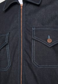 Mörk denimjacka med en central koppardragkedja, två bröstfickor med blå sömmar och en klassisk krage.