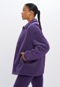Paarse pufferjack met een kraag, drukknoopsluiting, elastische manchetten en zijzakken. Glad textuur en een oversized pasvorm.