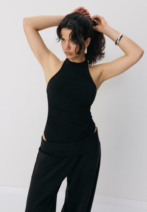 Donna con capelli ricci scuri che indossa una canotta nera e pantaloni neri, che si sistema i capelli con entrambe le mani alzate contro uno sfondo semplice.