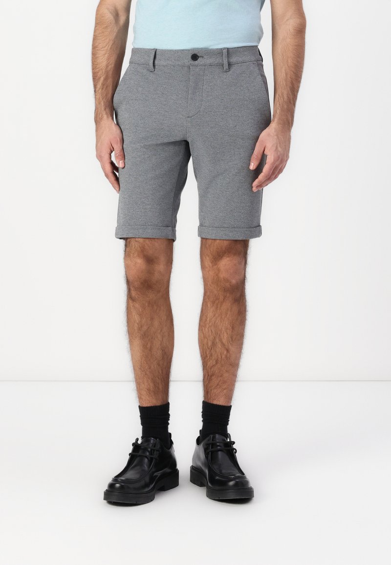 Grå strikkede shorts med rullekanter, med en enkelt knappelukking og beltehemper. Brukt med svarte lærsko og sokker.