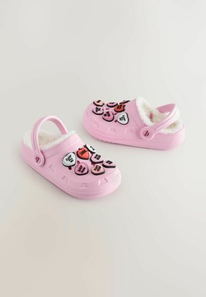 Sabots roses avec une doublure blanche duveteuse et des patches décoratifs en forme de cœur de différentes couleurs comportant du texte. Design à enfiler avec une sangle arrière.