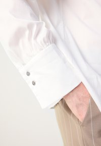Biała koszula z bufiastymi rękawami na mankietach, ozdobiona dwoma srebrnymi guzikami. Koszula ma gładką fakturę; zestawiona z beżowymi spodniami w paski.