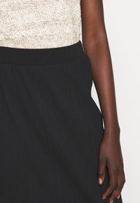 Jupe plissée noire avec une taille élastique. Le tissu a une texture lisse, créant une silhouette fluide. Au-dessus, un haut en tricot beige est visible.