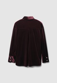 Camicia di velluto bordeaux con colletto, maniche lunghe e polsini con bottoni. Presenta una piega centrale sul retro per una maggiore libertà di movimento.