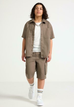 Jong persoon loopt, draagt een wit T-shirt, een bruin overhemd met korte mouwen en knopen, bijpassende cargoshorts, witte sneakers en gestreepte sokken.