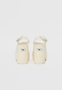 Sandales à plateforme de couleur crème avec une bride à la cheville, semelle texturée et détail du logo de marque à l'arrière. Finition en matériau lisse.