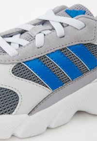 Zapatilla deportiva gris con acentos azules, que presenta una parte superior de malla y superposiciones de ante. Incluye cordones blancos y suela de goma texturizada.