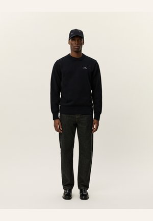 Svart genser, rund hals, rippeermansjetter, detaljer med logo. Kombinert med mørke jeans og svarte sko. Modell med svart caps, som står mot en lys bakgrunn.