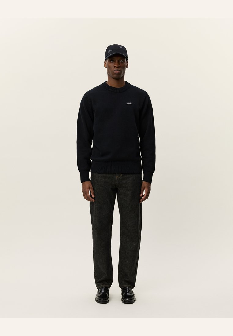 Maglione nero, collo tondo, polsini a coste, dettaglio con logo. Abbinato a jeans scuri e scarpe nere. Modello con berretto nero, in piedi contro uno sfondo chiaro.