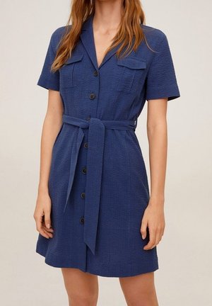 Nainen, joka pukeutunut lyhythihaiseen, tekstuuriin siniseen napitettuun mekkoon, jossa on kaulus, rintataskut ja solmittu vyö vyötäröllä.