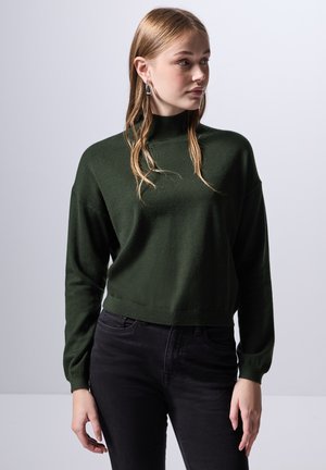 Suéter cortado de color verde oscuro, con cuello alto y mangas largas, hecho de una tela suave y texturizada. Combinado con jeans negros.