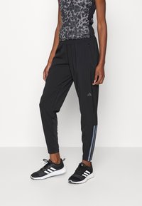 Svarta träningsbyxor med avsmalnande ben, som har en subtil glans, sidoficka med dragkedja och gråa ränder längs ytterseamna. Bärs med svarta sneakers.