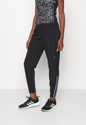 Zwarte sportbroek met taps toelopende pijpen, voorzien van een subtiele glans, een zijzak met rits en grijze strepen langs de buitennaad. G gedragen met zwarte sneakers.