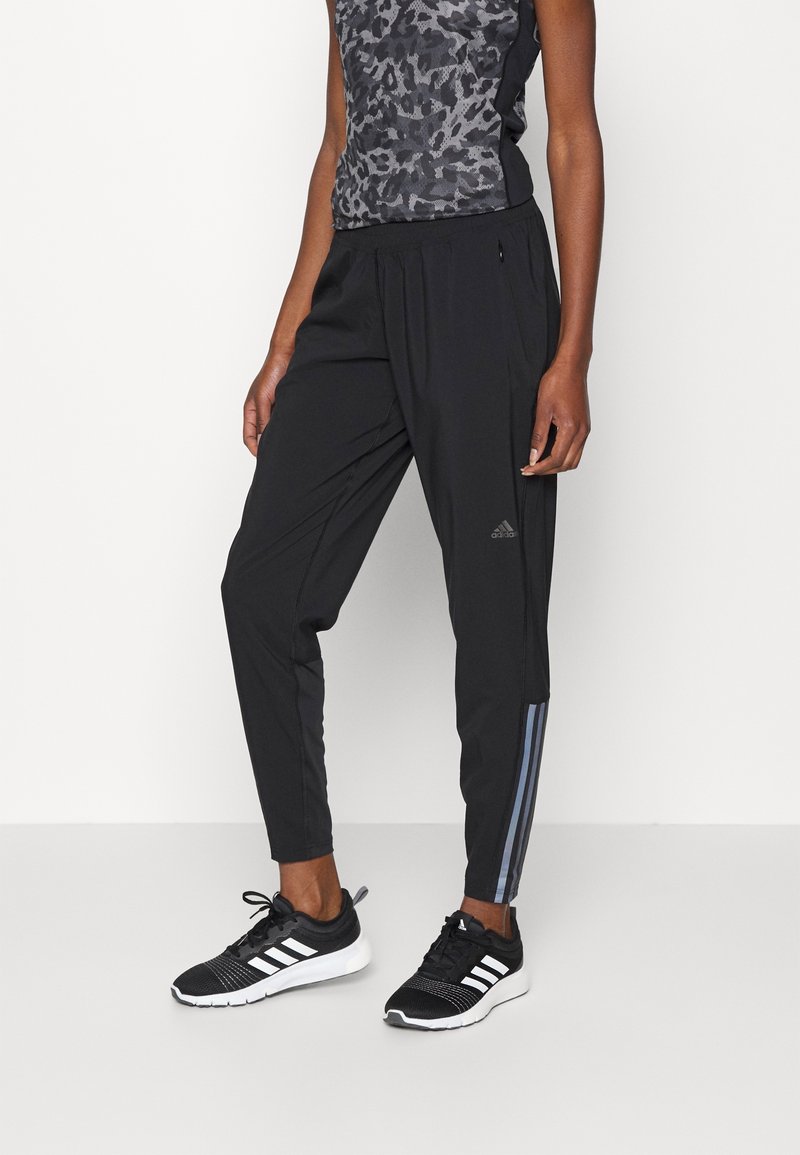 Svarta träningsbyxor med avsmalnande ben, som har en subtil glans, sidoficka med dragkedja och gråa ränder längs ytterseamna. Bärs med svarta sneakers.