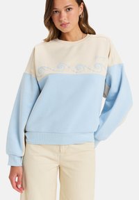 Sweat-shirt bleu clair et beige avec broderie de vagues sur la poitrine, poignets côtelés, coupe décontractée et en coton doux.