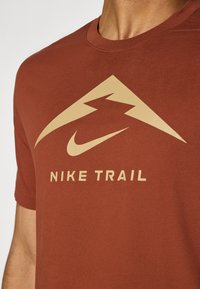 Rustfarget Nike Trail T-skjorte laget av bomull, med en beige fjell-logo og tekst nedenfor. Kort ermer med rund halsdesign.