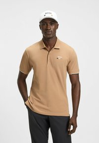 Tan polotröja med krage, med en logotyp på bröstet och kontrasterande trim på ärmarna. Matchad med svarta byxor.