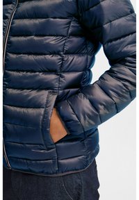 Jūrinės mėlynos puffer tipo striukės dizainas su kvadratuotas raštu, blizgiu paviršiumi ir šoniniais kišenėmis. Su priekiniais užtrauktukais ir elastinėmis rankovėmis, kad būtų patogiau.