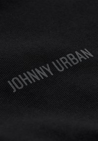 Černá textilie s embosovanými slovy "JOHNNY URBAN" v jemném šedém odstínu, s hladkou strukturou a jemným vzorem tkaní.