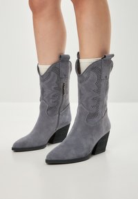 Botine din suede gri în stil cowboy, cu vârf ascuțit, decupaje decorative și toc pătrat. Are un fermoar pe lateral pentru a fi ușor de purtat.