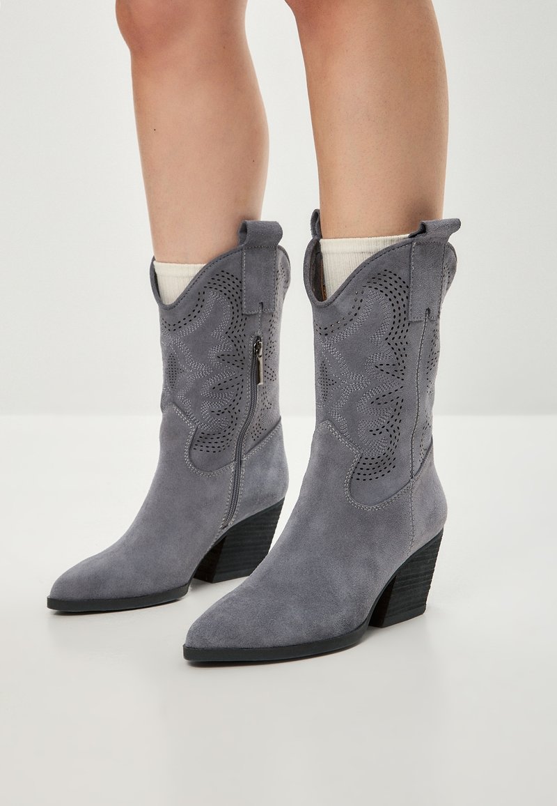 Botine din suede gri în stil cowboy, cu vârf ascuțit, decupaje decorative și toc pătrat. Are un fermoar pe lateral pentru a fi ușor de purtat.