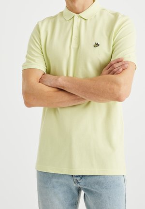 Mann mit verschränkten Armen, der ein hellgelbes Polohemd mit einem kleinen Vogel-Logo auf der Brust und eine hellblaue Jeans trägt.