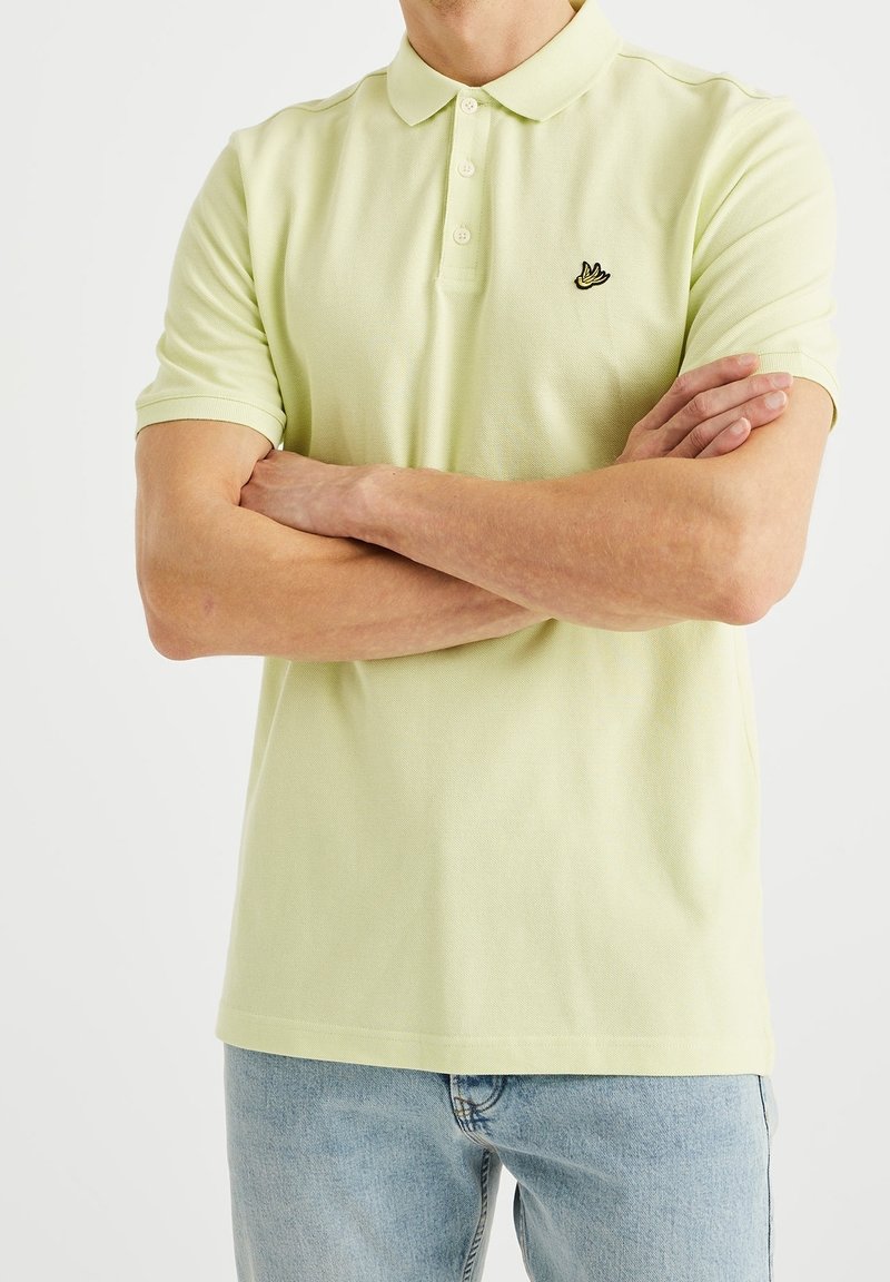 Homme les bras croisés portant un polo jaune clair avec un petit logo d'oiseau sur la poitrine et un jean bleu clair.