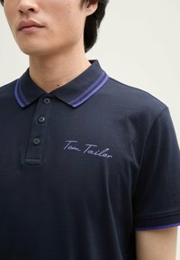 Sötétkék polo ing, két gombos nyakkal, lila csíkozott gallérral és hímzett "Tom Tailor" logóval a mellkason. Pamut anyagból.