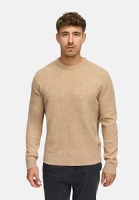 Beige gebreide trui met een ronde hals en geribbelde manchetten en zoom. De stof lijkt zacht en gestructureerd, met een klein merklabel aan de zijkant.
