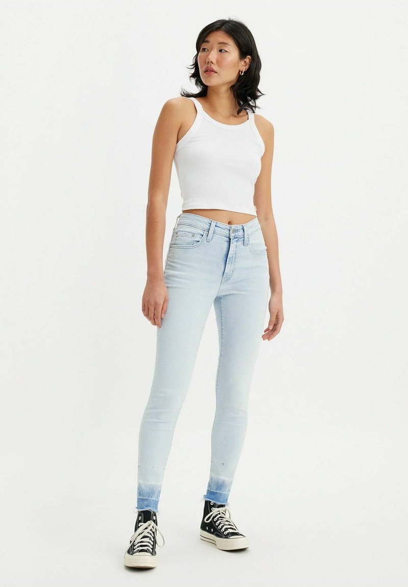 Helle blaue Skinny-Jeans mit einem verblassten Finish, hohe Taille und knöchellanger Schnitt, kombiniert mit einem weißen gerippten Tanktop und schwarzen Sneakers.