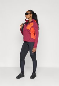 Bordeauxrode en oranje softshell jas met capuchon, gecombineerd met zwarte textuleggings en zwarte wandelschoenen, met een ritszak en details.
