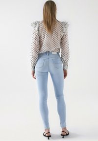 Jeans ajustados de color azul claro combinados con una blusa transparente de lunares que presenta mangas con volantes. Textura suave, cintura alta, silueta ajustada.