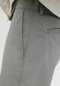 Pantaloni dintr-o țesătură ușoară, gri, cu o textură netedă, având un buzunar lateral și detalii de cusătură complexe.