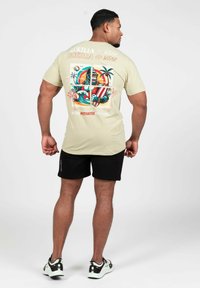 Beige T-shirt met korte mouwen met een kleurrijke gorilla-print op de rug, zwarte shorts en meerkleurige sneakers. Soepele textuur, relaxte pasvorm.