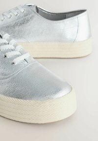 Baskets basses métalliques argentées avec des lacets blancs et des semelles en caoutchouc texturées épaisses de couleur crème, vues sous un angle de trois quarts.