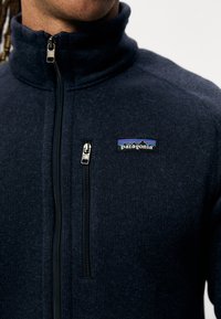Veste polaire marine avec un col haut, dotée d'une poche frontale zippée, de zips noirs et d'un logo Patagonia sur le côté supérieur gauche.