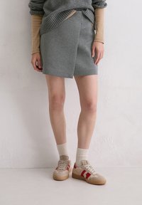 Grauer Wickelrock mit glatter Textur, kombiniert mit beigen Turnschuhen mit roten Streifen und strukturierten Details, ergänzt durch cremefarbene Socken.