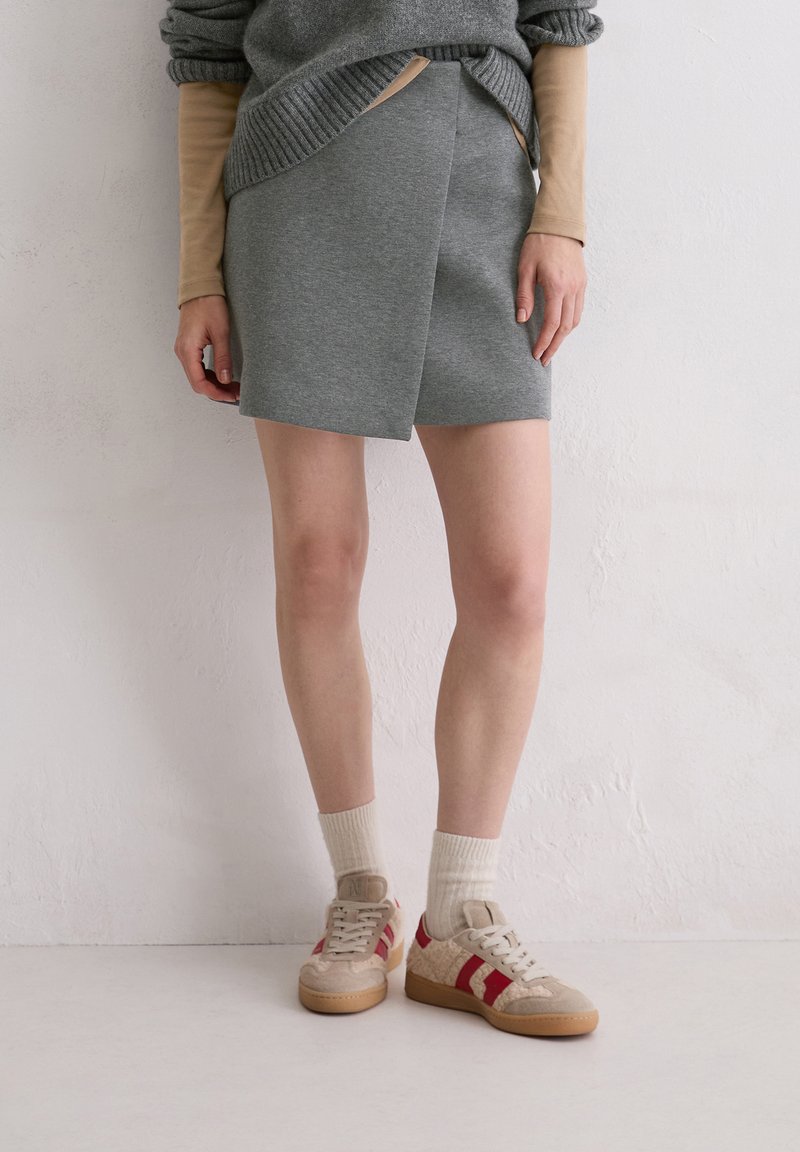 Grauer Wickelrock mit glatter Textur, kombiniert mit beigen Turnschuhen mit roten Streifen und strukturierten Details, ergänzt durch cremefarbene Socken.